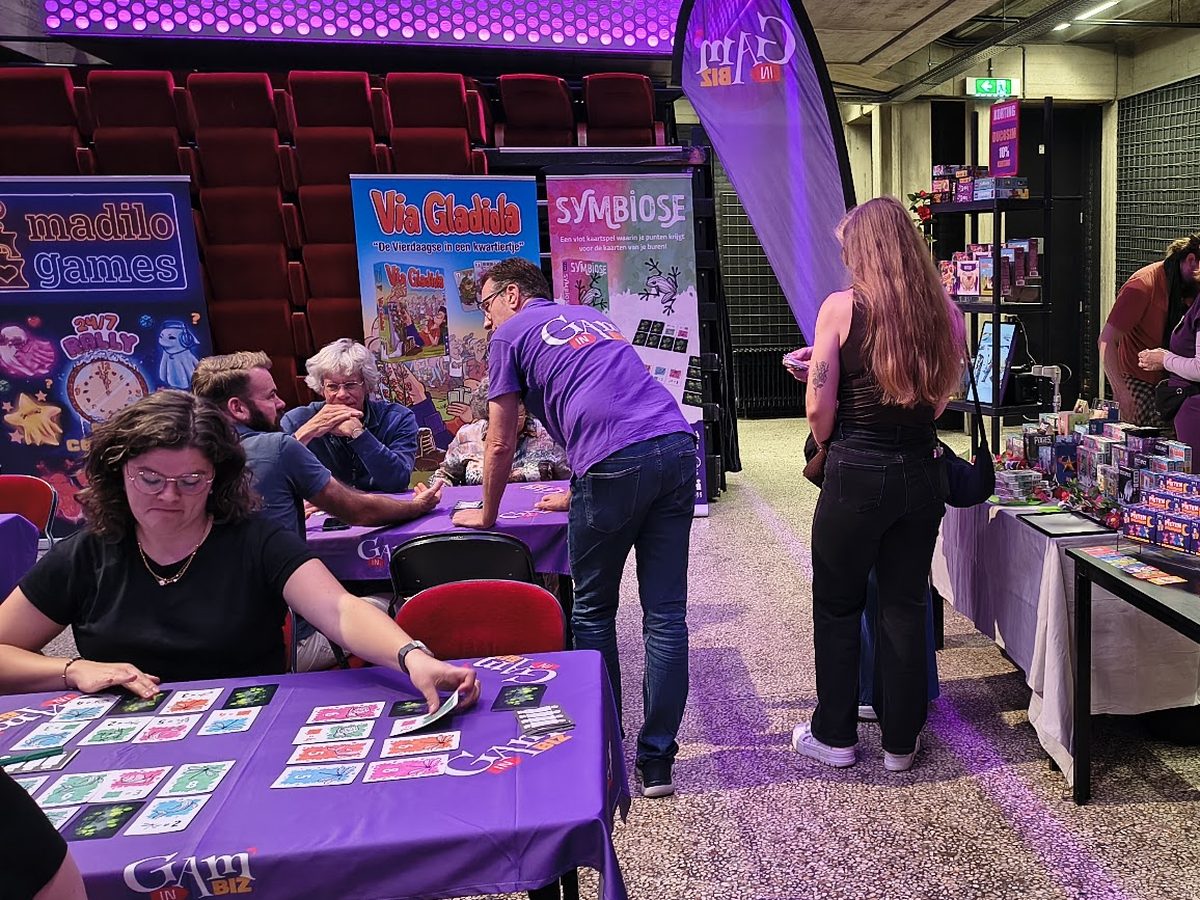 Het Gam’inBIZ-demoteam op een spellenbeurs: bezoekers spelen kaartspellen aan paarse Gam’inBIZ-tafels terwijl een demoteamlid in een paars shirt uitleg geeft